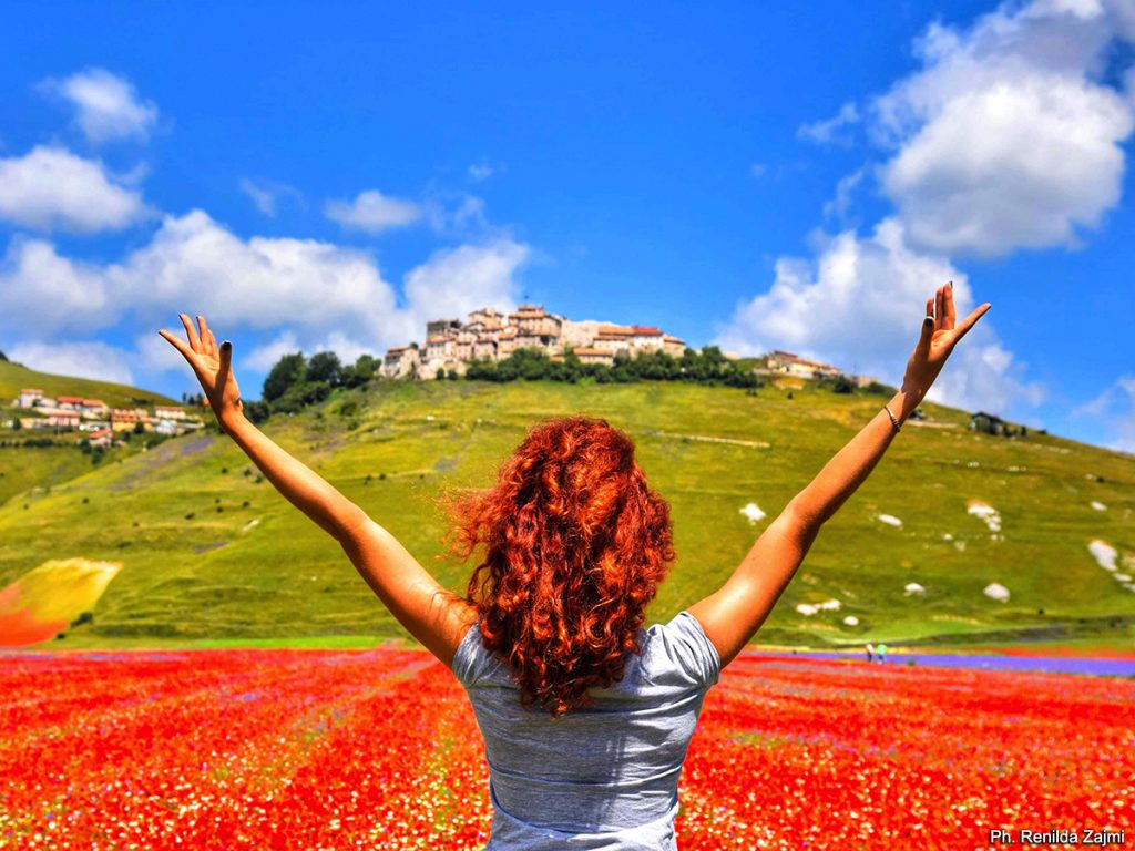 Una ragazza di spalle davanti alla piana di Castelluccio fiorita con il borgo sullo sfondo.