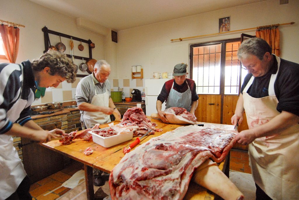 quattro persone attorno a un tavolo di legno intente a separare le varie parti del maiale macellato per ricavarne salumi e altri prodotti.